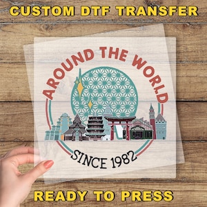 May include: A clear DTF transfer sheet featuring a colorful design with the text "AROUND THE WORLD SINCE 1982". The design includes illustrations of various landmarks and a geometric sphere. The text "READY TO PRESS" is also visible.