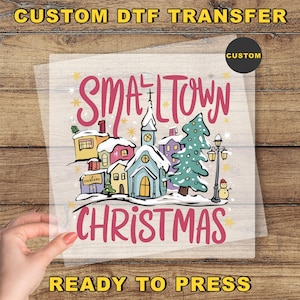 May include: A DTF transfer with a winter village scene. The design features a church, houses, and a Christmas tree, with the words "Smalltown Christmas" in pink. The words "Custom DTF Transfer" and "Ready to Press" are also visible.
