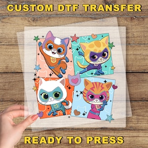 May include: A clear sheet featuring a DTF transfer design with four cartoon superhero cats in vibrant colors. Each cat is in a different pose and costume, surrounded by stars and hearts. The text "CUSTOM DTF TRANSFER" and "READY TO PRESS" are at the top and bottom.