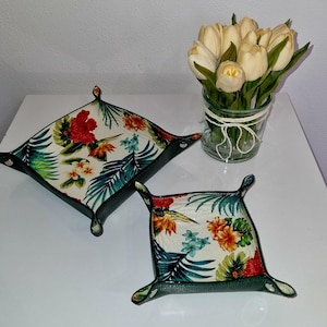May include: Two square decorative trays with a tropical floral pattern featuring red hibiscus flowers, green leaves, and blue accents. The trays have a dark green border and are placed on a white surface. A vase of white tulips is in the background.