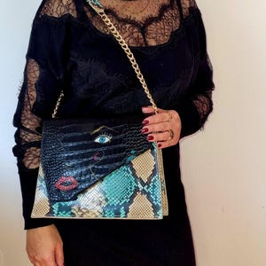 Könnte beinhalten: Schwarze und mehrfarbige Clutch mit einem goldenen Kettenriemen. Die Clutch hat eine schwarze, krokodiltexturierte Klappe mit einem Augen- und Lippen-Design. Der untere Teil hat ein Schlangenmuster in Türkis, Beige und Schwarz.