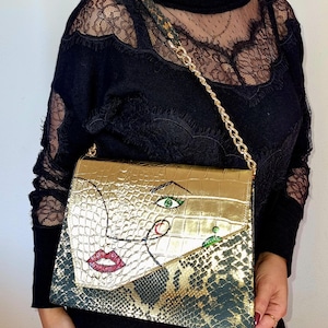 Op de afbeelding: Goudkleurige handtas met een kettingriem. De tas heeft een klep met krokodillenstructuur met een geschilderd gezichtsontwerp, inclusief rode lippen en een groen oog. Het onderste gedeelte heeft een slangenpatroon.