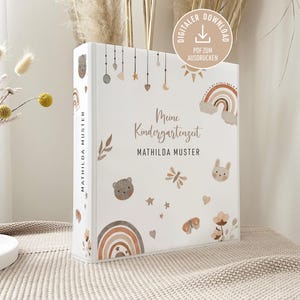 May include: A white binder with the text "Meine Kindergartenzeit" and "MATHILDA MUSTER" in brown. The binder is decorated with illustrations of a rainbow, bear, bunny, butterfly, and stars. A digital download icon is in the upper right corner.