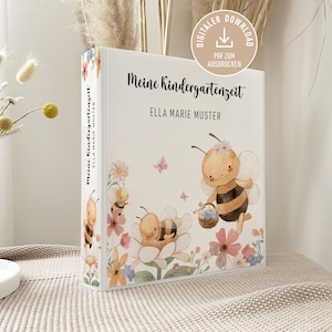 Puede incluir: Una carpeta blanca titulada Meine Kindergartenzeit con el nombre Ella Marie Muster impreso en la parte frontal. La portada presenta ilustraciones en acuarela de abejas, flores y mariposas. Un sello circular indica una descarga digital en PDF.