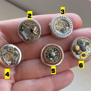May include: Six small round silver-toned metal objects with intricate clockwork designs. Each object has a different design and is numbered 1 through 6.