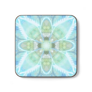 May include: A square coaster with rounded corners featuring a symmetrical, abstract pattern in shades of light blue, pale green, and white. The design consists of radiating, curved lines and soft, glowing circular shapes centered on the surface.