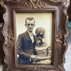 May include: A sepia-toned framed photograph featuring a man in a suit and a person with a skeletal head and goggles. The ornate, dark brown frame has floral and scrollwork details, adding a vintage aesthetic to the image.