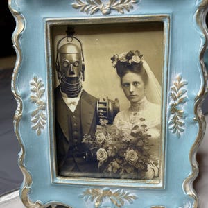 May include: A vintage-style framed photograph featuring a sepia-toned image of a bride and a robot groom. The frame is light blue with gold accents. The image shows the couple holding a bouquet of roses.