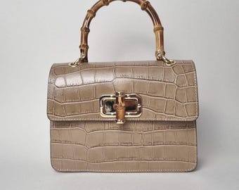 May include: A taupe-colored handbag with a crocodile-skin texture. The bag features a bamboo handle and a gold-toned clasp. The rectangular bag has a flap closure and a structured design, suitable for everyday use.