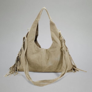 May include: A beige suede shoulder bag with a long adjustable strap and fringe detailing. The bag has a curved shape and a stitched design down the center. The bag has gold-colored hardware.