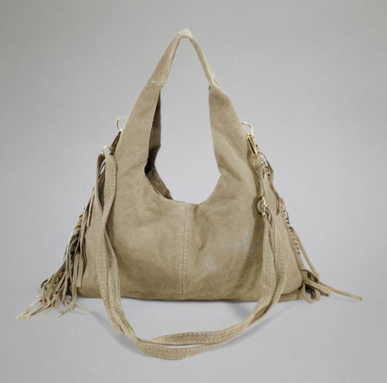 May include: A beige suede shoulder bag with a long adjustable strap and fringe detailing. The bag has a curved shape and a stitched design down the center. The bag has gold-colored hardware.