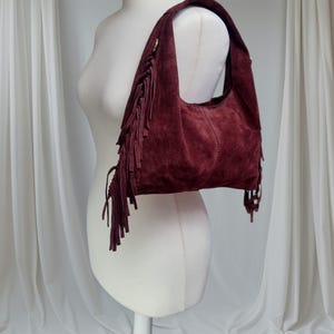 May include: A burgundy suede shoulder bag with fringe detailing. The bag is displayed on a white mannequin, showcasing its curved shape and the texture of the suede. The fringe hangs from the sides, adding a bohemian flair.