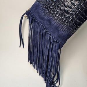 Puede incluir: Un bolso azul marino con un cuerpo de cuero con textura de cocodrilo y detalles de flecos de ante. El bolso presenta una larga franja a lo largo del borde inferior y un panel de ante en la parte superior. El bolso es de color azul oscuro.