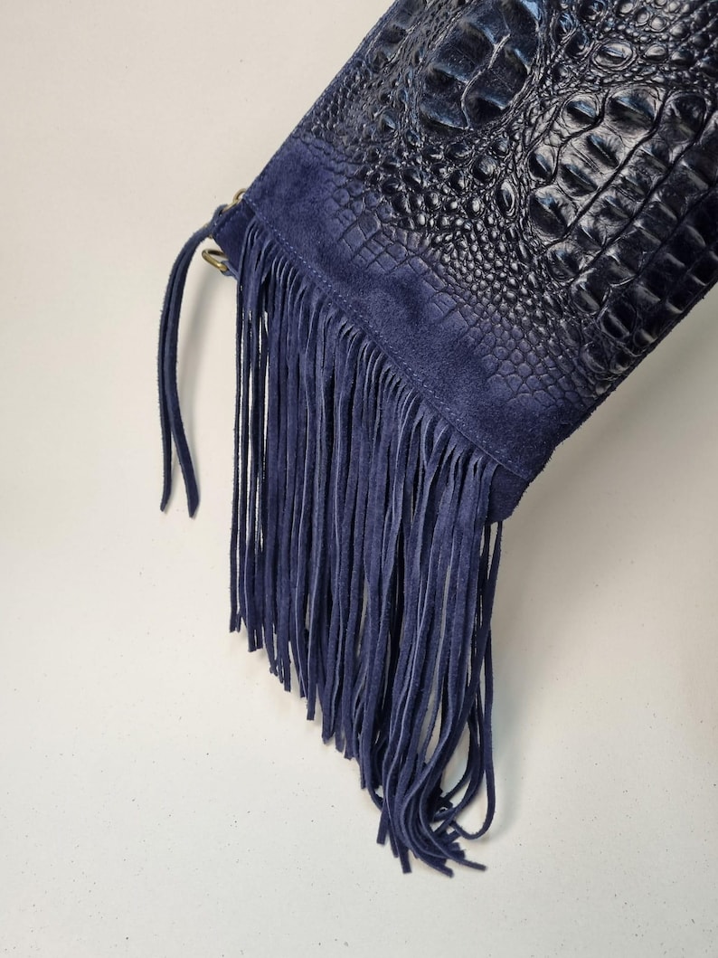 Puede incluir: Un bolso azul marino con un cuerpo de cuero con textura de cocodrilo y detalles de flecos de ante. El bolso presenta una larga franja a lo largo del borde inferior y un panel de ante en la parte superior. El bolso es de color azul oscuro.