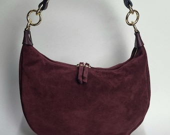 May include: A burgundy suede crescent-shaped handbag with a top handle and gold-tone hardware. The bag features a zipper closure and a dark purple leather handle. The bag is a stylish accessory.