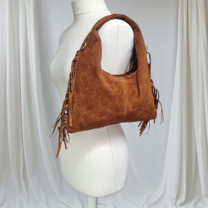 May include: A brown suede shoulder bag with a rounded shape and a single handle. The bag features decorative fringe along the sides and bottom. The bag is displayed on a white mannequin.