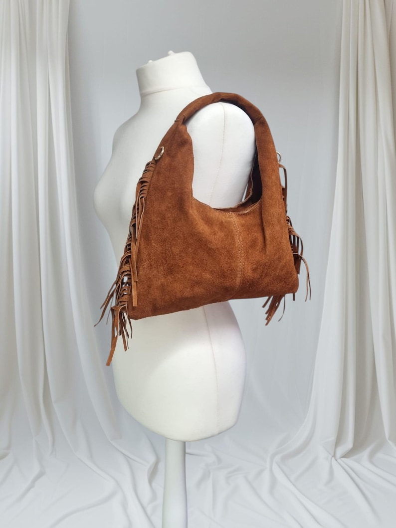 May include: A brown suede shoulder bag with a rounded shape and a single handle. The bag features decorative fringe along the sides and bottom. The bag is displayed on a white mannequin.