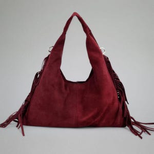 May include: A burgundy suede hobo bag with long fringe detailing. The bag has a single shoulder strap and a spacious interior. The fringe is along the sides of the bag. The bag is a deep red color.