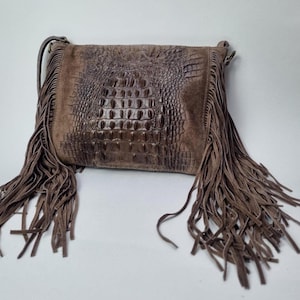 May include: A brown leather crossbody bag with a crocodile-skin effect on the front. The bag has long, flowing fringe on the sides and a shoulder strap. The bag is a medium brown colour.