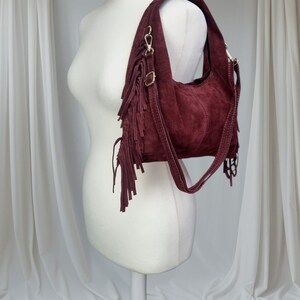 May include: A burgundy suede shoulder bag with fringe detailing. The bag features a curved shape, a shoulder strap, and gold-colored hardware. The fringe hangs from the front of the bag, adding a bohemian flair. The bag is displayed on a mannequin.