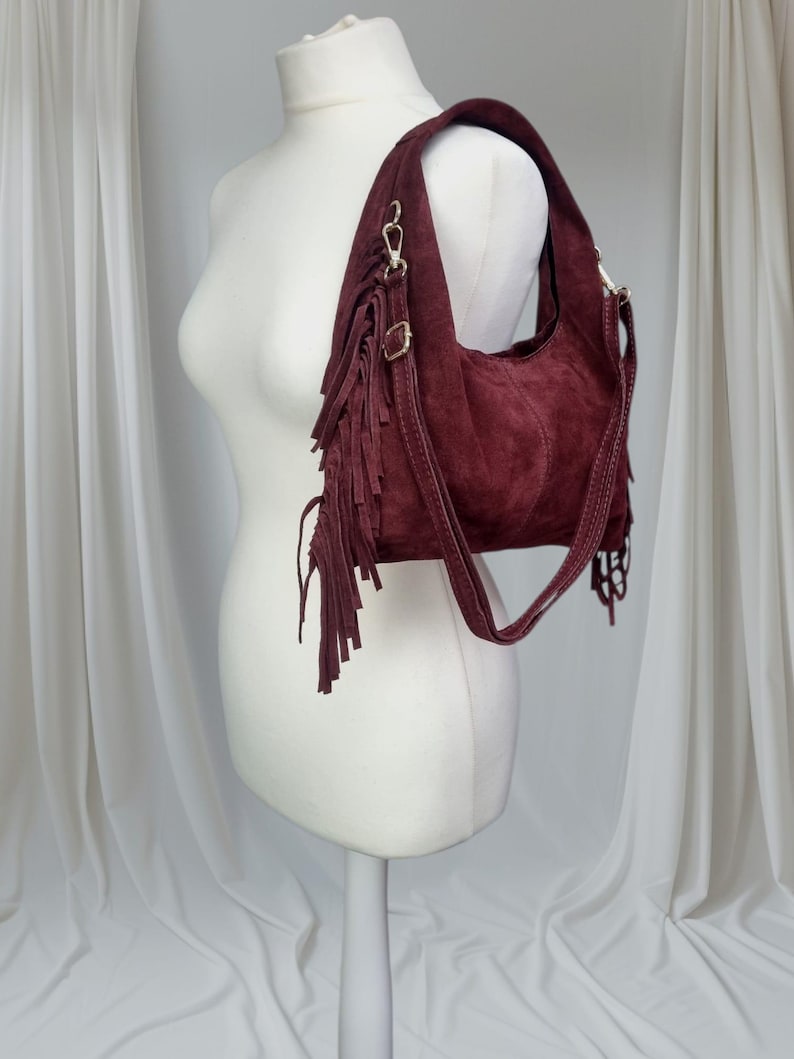 May include: A burgundy suede shoulder bag with fringe detailing. The bag features a curved shape, a shoulder strap, and gold-colored hardware. The fringe hangs from the front of the bag, adding a bohemian flair. The bag is displayed on a mannequin.