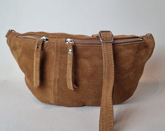 May include: A brown suede fanny pack with two zippered compartments and a long adjustable strap. The bag has a curved shape and a silver-colored clasp. The zippers have leather pull tabs.