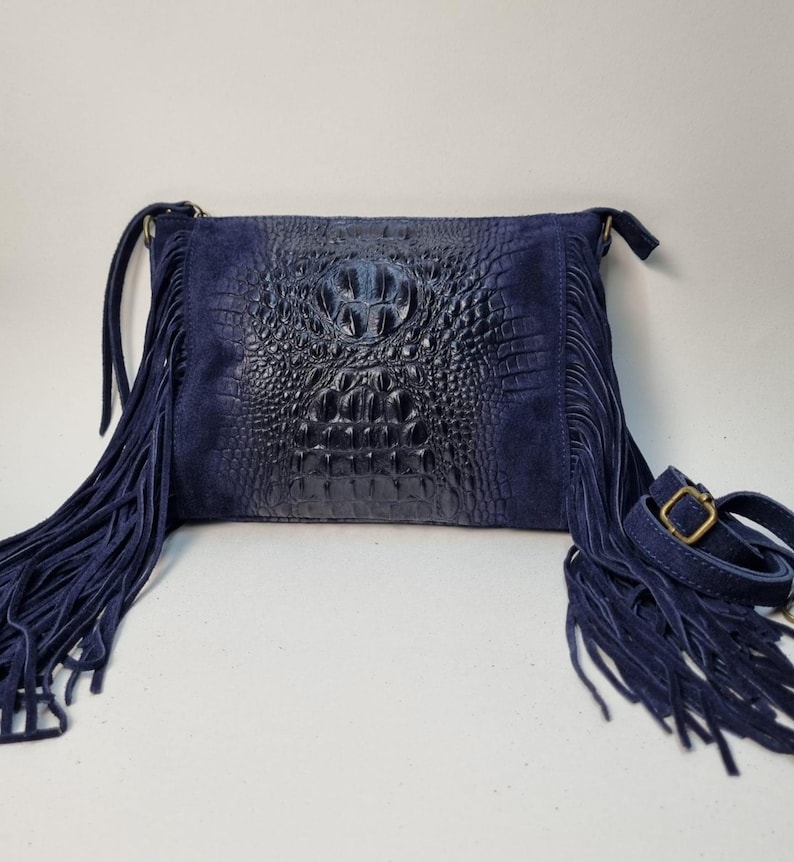 Puede incluir: Bolso bandolera azul marino con un panel central con relieve de cocodrilo y flecos de ante. El bolso tiene una correa ajustable y una hebilla met&aacute;lica. El bolso mide aproximadamente 25 cm de ancho y 20 cm de alto.