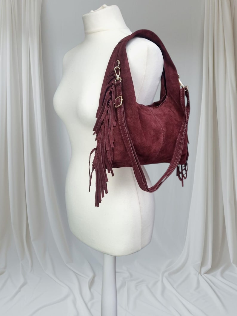 Op de afbeelding: Een bordeauxrode su&egrave;de schoudertas met franjes. De tas heeft een gebogen vorm, een schouderriem en goudkleurige hardware. De franjes hangen aan de zijkant van de tas en geven een bohemien uitstraling. De tas wordt op een mannequin getoond.