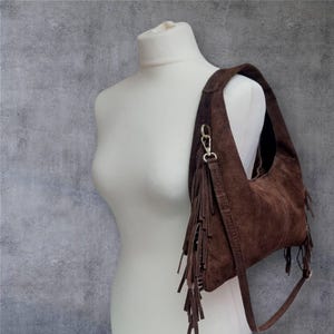 May include: A brown suede shoulder bag with fringe detailing. The bag is displayed on a white mannequin against a gray background. The bag has a long strap and a gold-colored clasp.