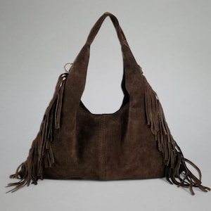 May include: A brown suede shoulder bag with long fringe detailing. The bag has a large, open interior and long straps. The fringe hangs from the sides and bottom of the bag, adding a bohemian flair. The bag is made of a soft, suede material.