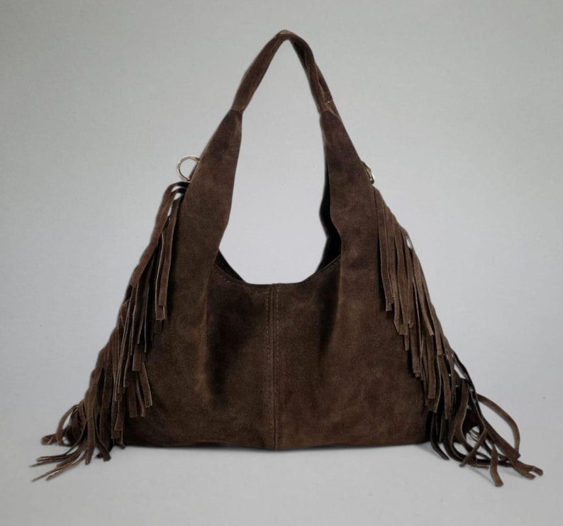 May include: A brown suede shoulder bag with long fringe detailing. The bag has a large, open interior and long straps. The fringe hangs from the sides and bottom of the bag, adding a bohemian flair. The bag is made of a soft, suede material.