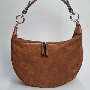 May include: A brown suede crescent-shaped handbag with a zip closure. The bag features a brown leather handle attached to gold-coloured rings. The bag is a warm, earthy tone, suitable for everyday use.