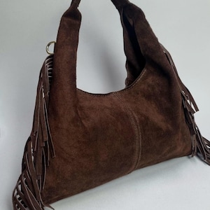 May include: A brown suede shoulder bag with a single handle. The bag features long fringe detailing along the sides and a curved shape. The bag is made of a soft, textured material.