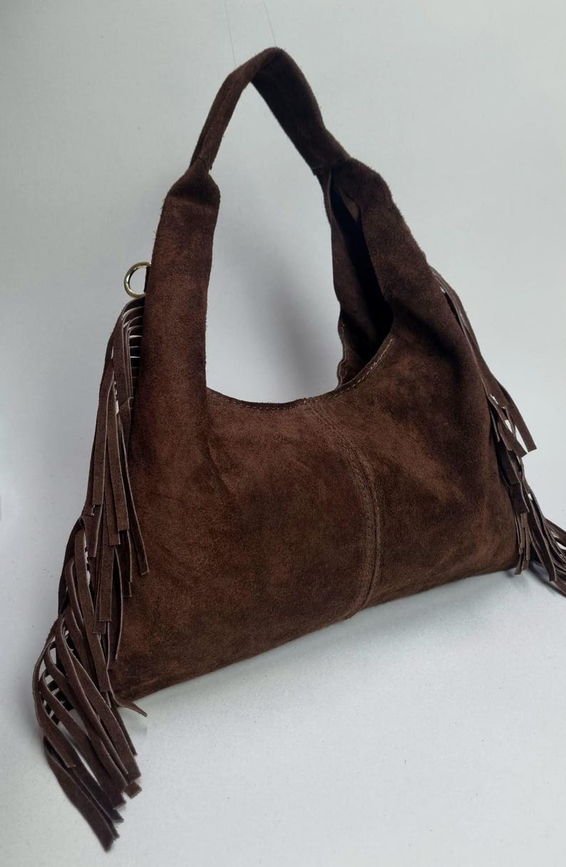 May include: A brown suede shoulder bag with a single handle. The bag features long fringe detailing along the sides and a curved shape. The bag is made of a soft, textured material.