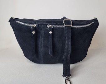 May include: A dark navy blue suede fanny pack with silver zippers and hardware. The bag has a long adjustable strap with a buckle and a metal clasp. The fanny pack has three zippered compartments.
