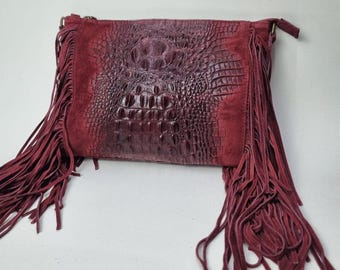 May include: A burgundy leather crossbody bag with a crocodile-embossed flap and suede fringe. The bag features a zipper closure and a long, adjustable strap. The bag's color is a deep, rich shade of red.