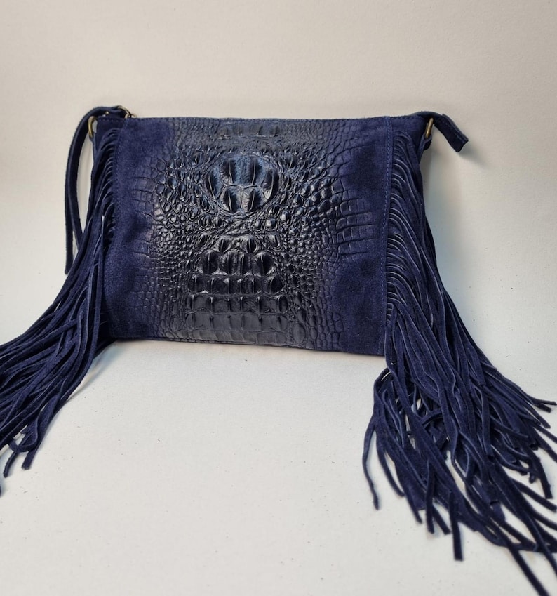 Puede incluir: Un bolso bandolera de ante y cuero azul marino con flecos. El bolso presenta un panel central de cuero con relieve de cocodrilo y laterales de ante. El bolso tiene forma rectangular y una correa larga.