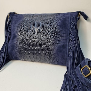 Puede incluir: Un bolso bandolera azul marino con un panel central en relieve de cocodrilo y paneles laterales de ante. El bolso presenta largas flecos en los laterales y una correa ajustable con una hebilla dorada.