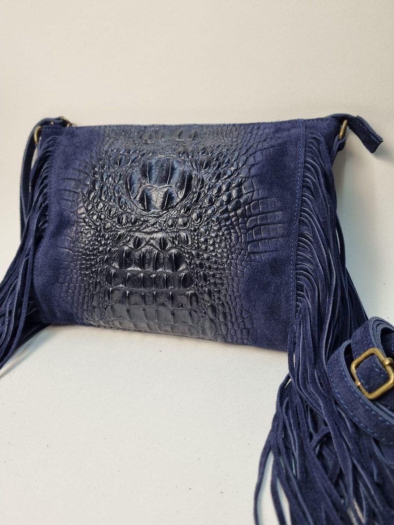Puede incluir: Un bolso bandolera azul marino con un panel central en relieve de cocodrilo y paneles laterales de ante. El bolso presenta largas flecos en los laterales y una correa ajustable con una hebilla dorada.