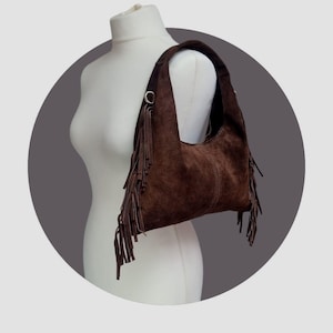 May include: A brown suede shoulder bag with a curved shape and long fringe detailing. The bag has a single shoulder strap and metal ring accents. The bag is displayed on a white mannequin.