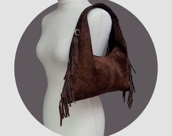 May include: A brown suede shoulder bag with a curved shape and long fringe detailing. The bag has a single shoulder strap and metal ring accents. The bag is displayed on a white mannequin.