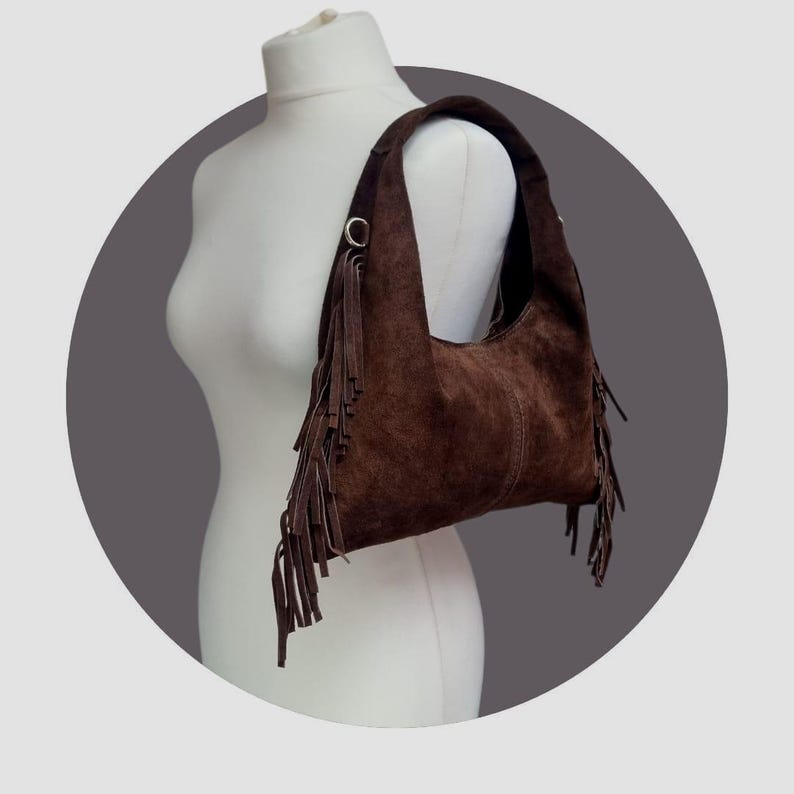 May include: A brown suede shoulder bag with a curved shape and long fringe detailing. The bag has a single shoulder strap and metal ring accents. The bag is displayed on a white mannequin.