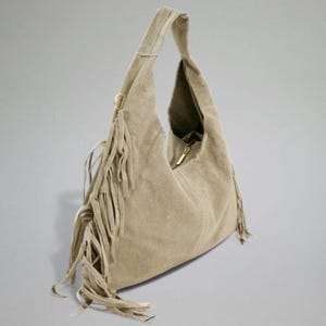 May include: A beige suede hobo bag with a single shoulder strap. The bag features a zipper closure and decorative fringe along the side. The bag is a neutral color and has a soft, textured appearance.