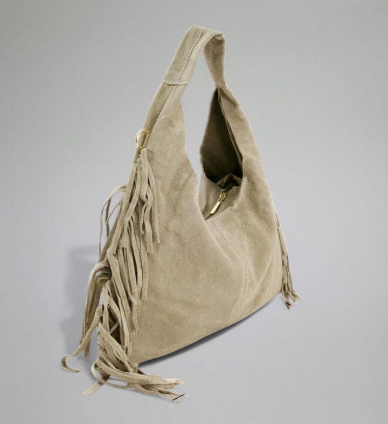May include: A beige suede hobo bag with a single shoulder strap. The bag features a zipper closure and decorative fringe along the side. The bag is a neutral color and has a soft, textured appearance.