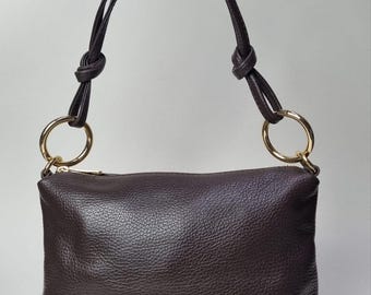 May include: A dark brown leather handbag with a rectangular shape. The bag features a zip closure and a handle made of the same leather, attached with gold-colored rings. The handle is knotted at the top.
