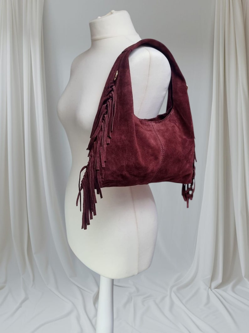Op de afbeelding: Een bordeauxrode su&egrave;de hobo-tas met franjedetails. De tas heeft een afgeronde vorm en een enkele schouderriem. De franjes hangen aan de zijkanten en onderkant van de tas, wat een bohemien uitstraling geeft. De tas wordt getoond op een witte mannequin.