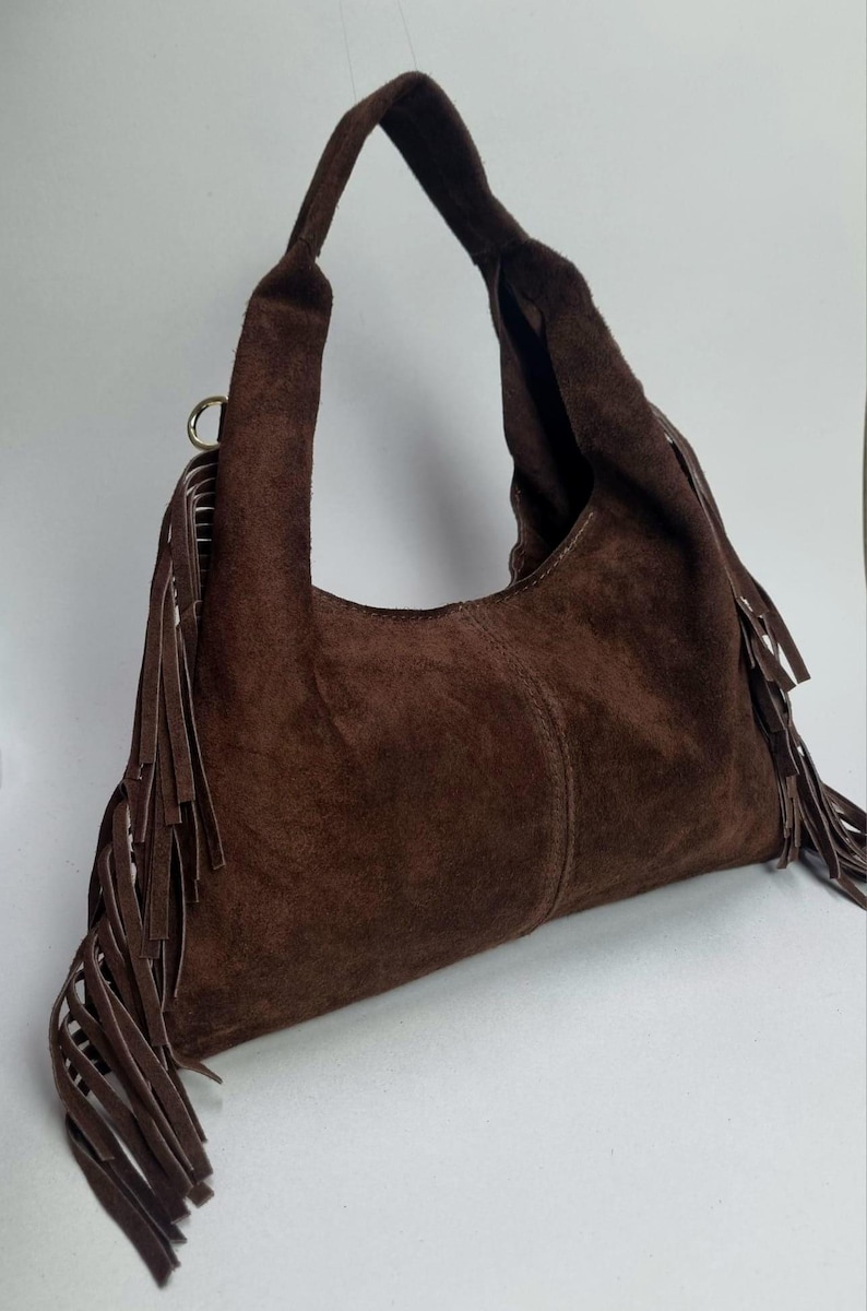 Op de afbeelding: Een bruine su&egrave;de schoudertas met een gebogen vorm en een enkele handgreep. De tas heeft lange franjes langs de zijkanten en een goudkleurige ringbevestiging voor de handgreep.