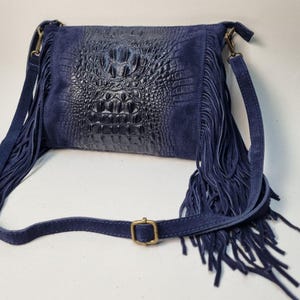 Puede incluir: Un bolso bandolera azul marino con un panel central texturizado con forma de cocodrilo. El bolso presenta flecos de ante en los laterales y una larga correa ajustable. Una hebilla dorada asegura la correa.