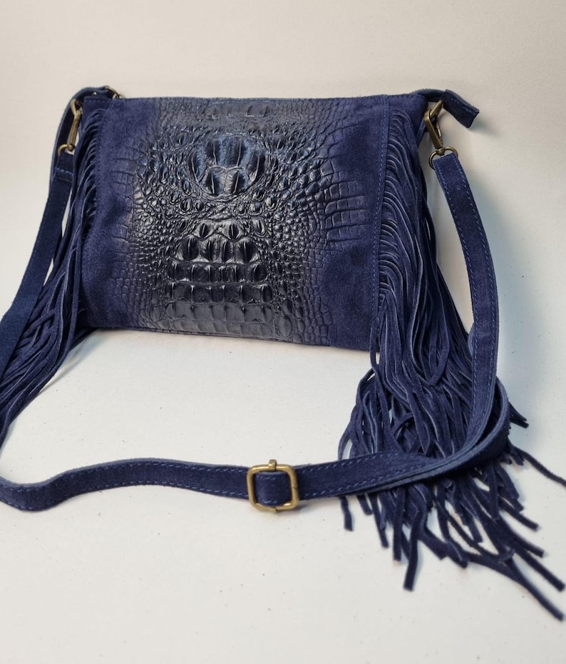 Puede incluir: Un bolso bandolera azul marino con un panel central texturizado con forma de cocodrilo. El bolso presenta flecos de ante en los laterales y una larga correa ajustable. Una hebilla dorada asegura la correa.