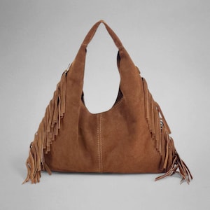 May include: A brown suede hobo bag with long fringe detailing. The bag has a curved shape with a single shoulder strap and visible stitching down the center. The fringe is along the sides and bottom of the bag.